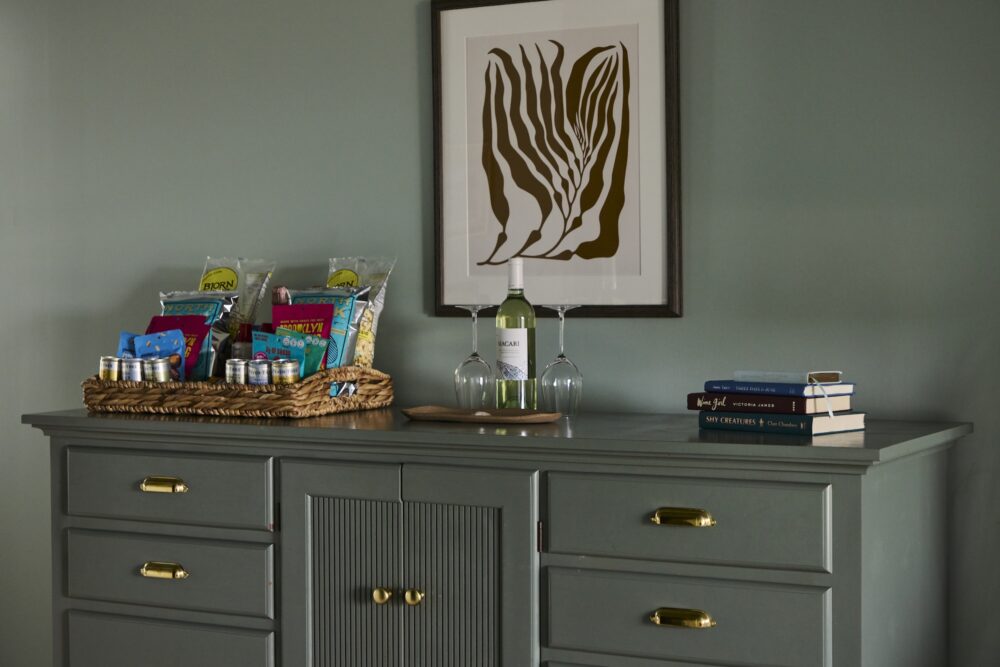 A dresser in a guest room with amenities, wine, and books.