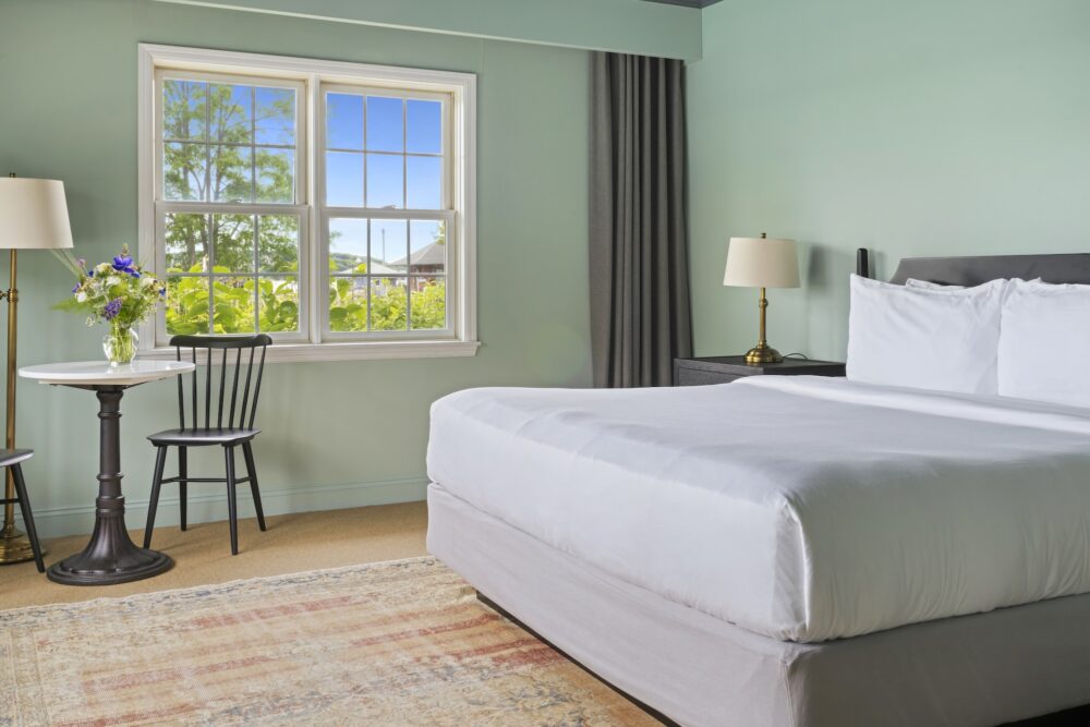 A guest bedroom with a queen bed, and table in the corner with a vase of flowers.