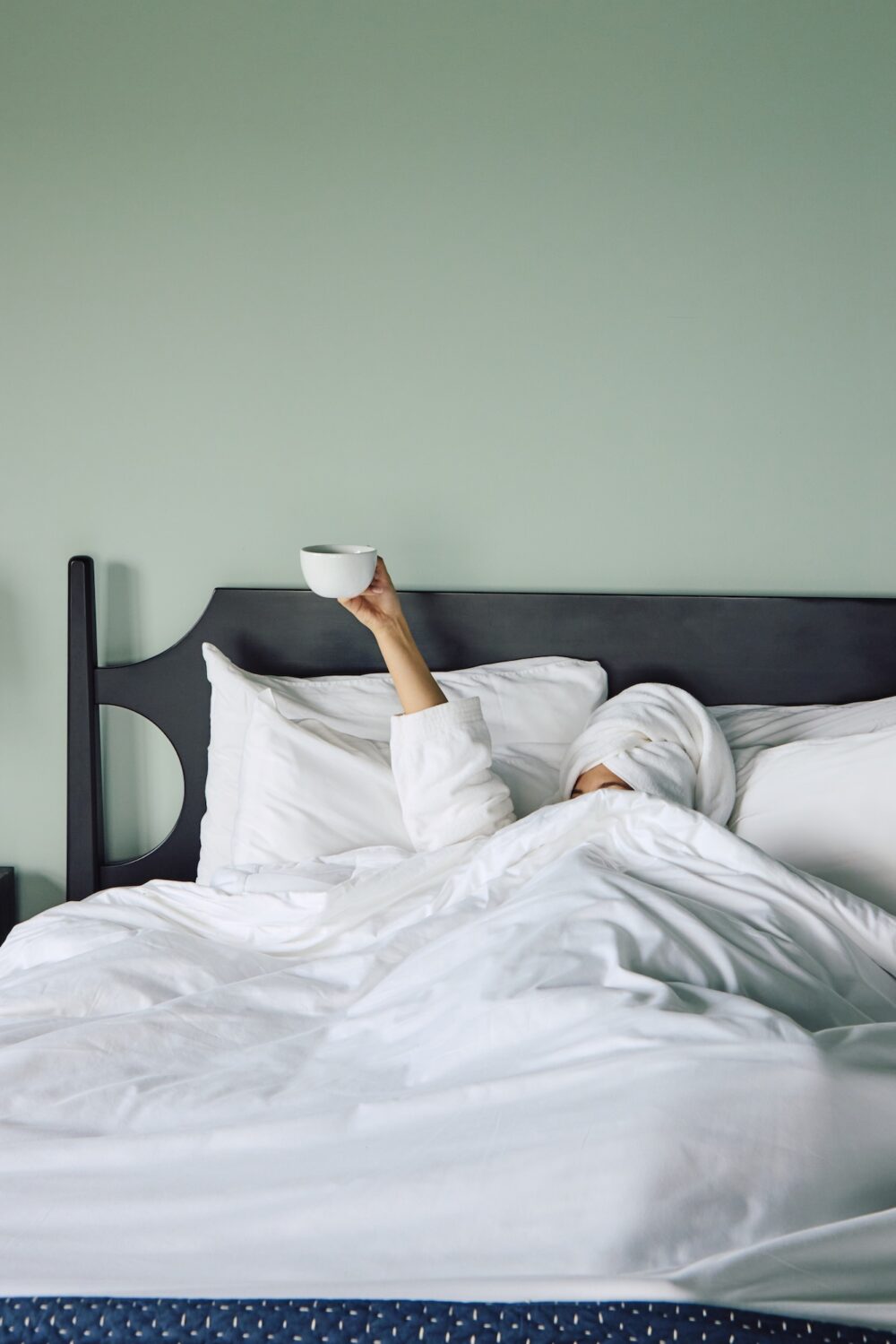 A woman in bed, under the covers holding up a cup of coffee.