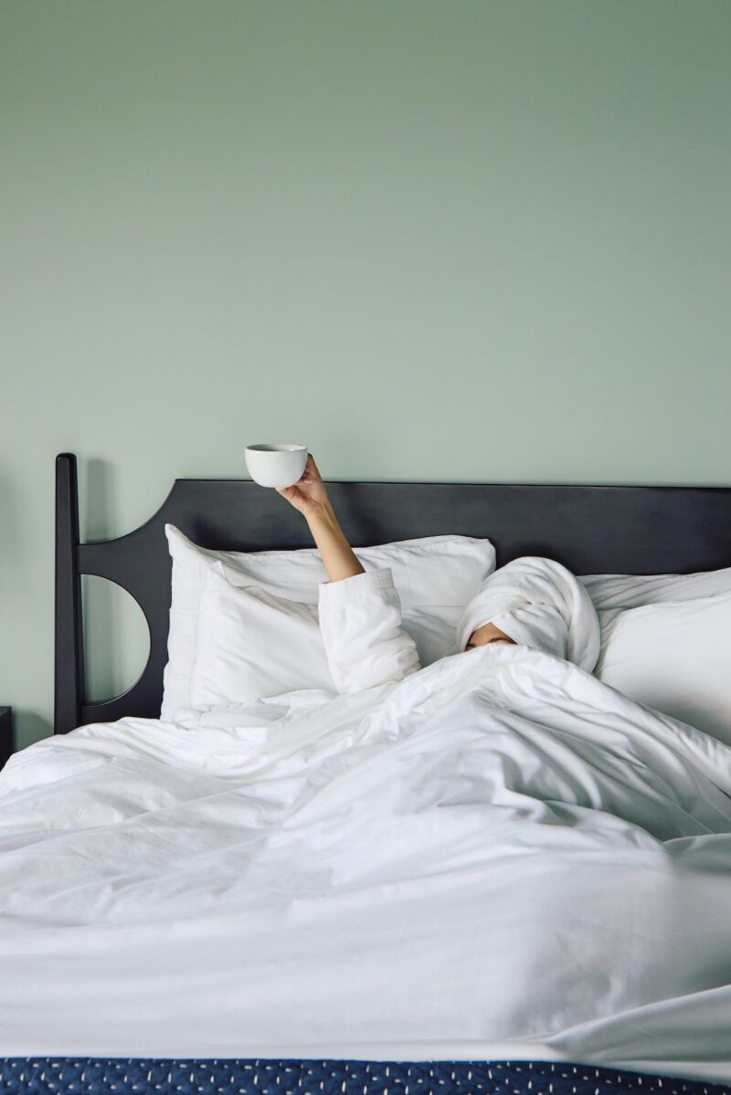 A woman in bed, under the covers holding up a cup of coffee.