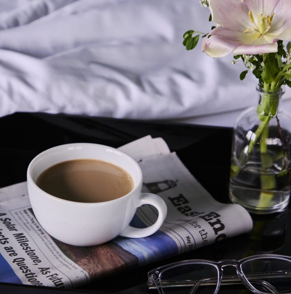 A cup of coffee on a night stand with a small vase of flowers.