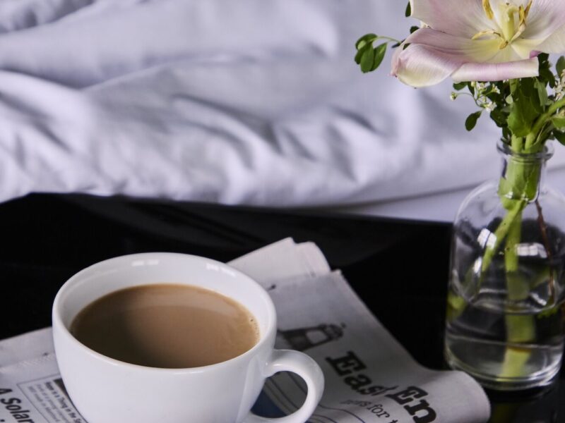 A cup of coffee on a night stand with a small vase of flowers.